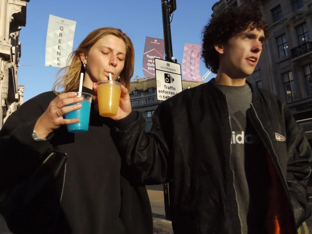A woman and a man standing on a city street, each holding a colorful drink with a straw. The woman drinks from a blue cup, and the man holds a yellow one. The woman has long blonde hair and earrings, and the man has curly dark hair and is wearing a b