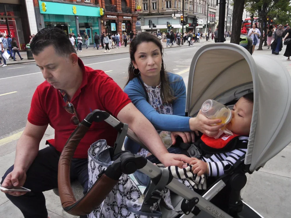 A family sits on a bench at a city street with a man, woman, and a baby in a stroller; the woman is feeding the baby with a bottle while the man looks at his phone. Background shows busy urban scene with pedestrians, shops, and traffic.