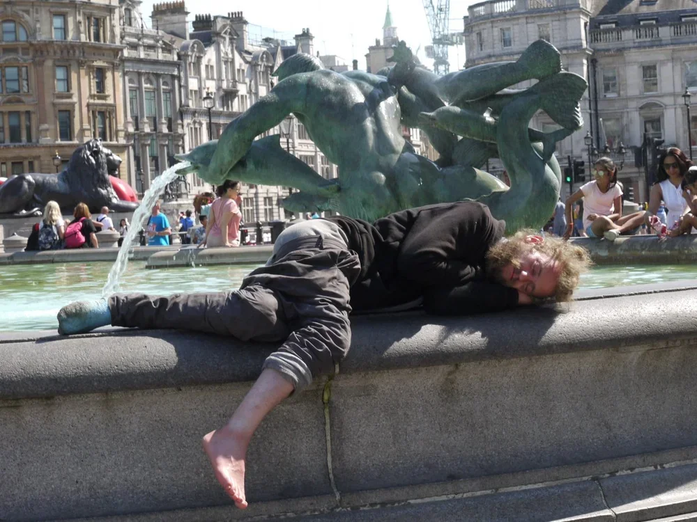 A person lying on the edge of a fountain in a city square, resting with their head on the fountain ledge. The background shows fountain sculptures and buildings with classical architecture, and several people sitting or walking nearby.