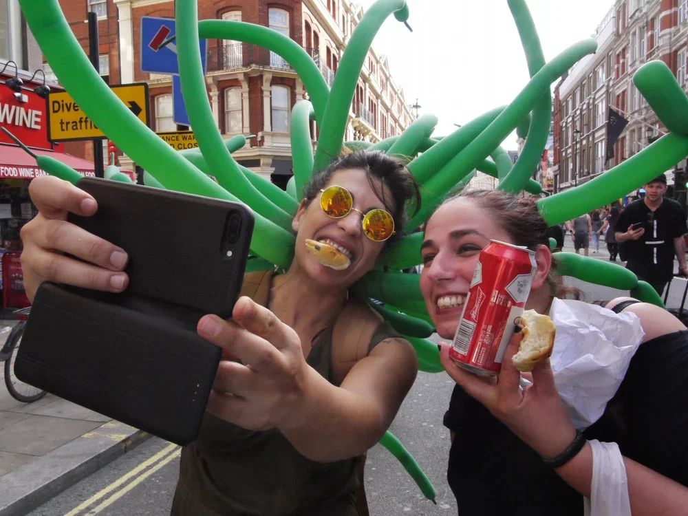Two women wearing inflatable green devil horns, taking a selfie with a smartphone. One is holding a donut and a soda can, and both are smiling.