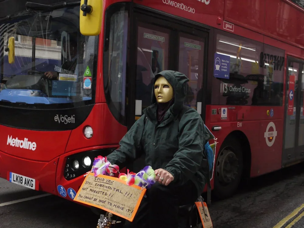 A person wearing a mannequin mask and a dark hooded jacket riding a bicycle with a basket filled with colorful flowers, in front of a red London bus.