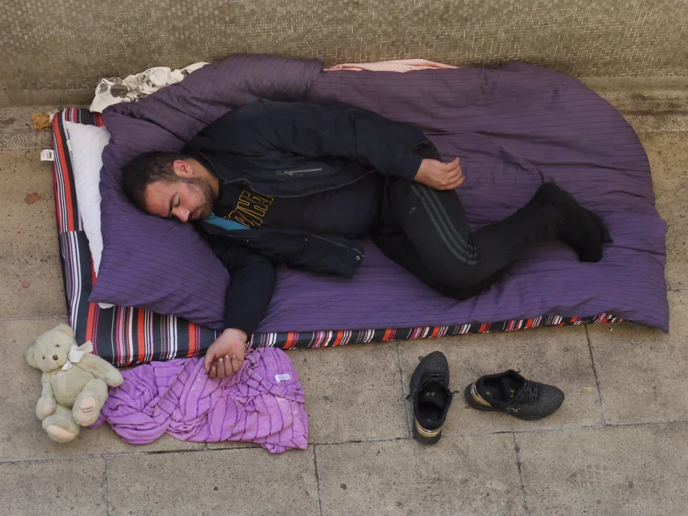 A man sleeping on a purple mattress outside on a sidewalk, with a teddy bear and a purple towel beside him, and his black shoes placed nearby.