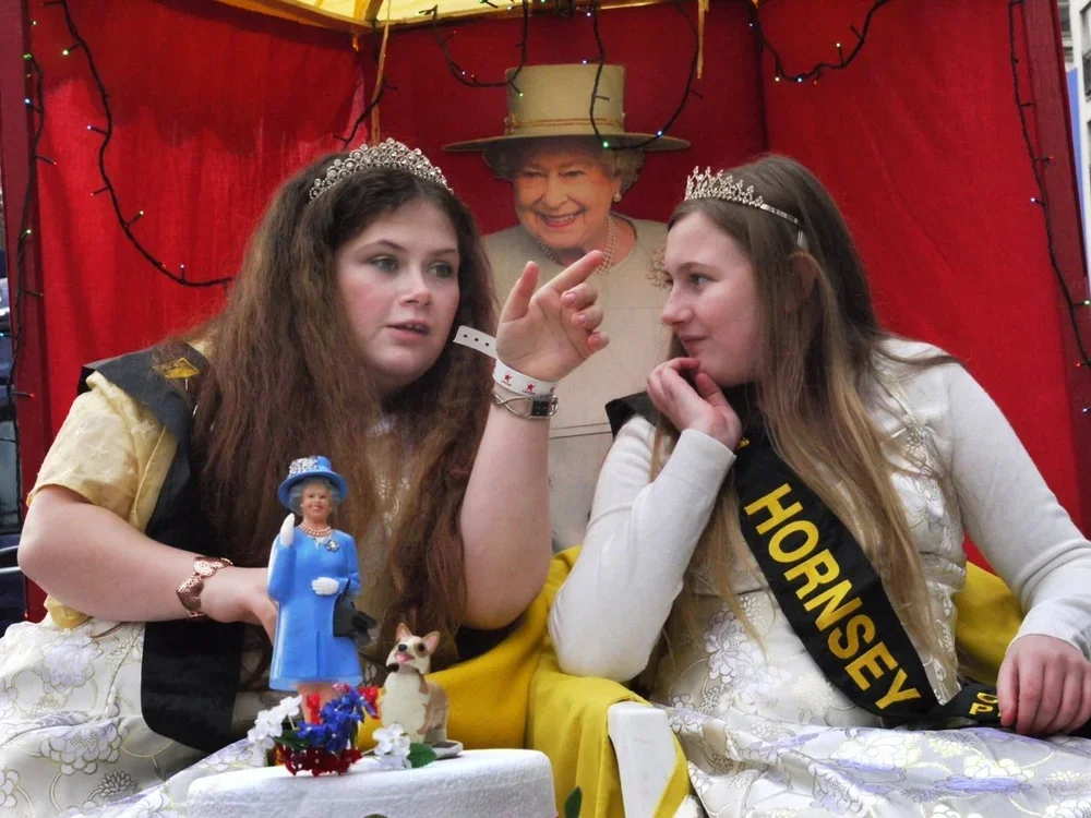 Three women dressed as queens with crowns, including Queen Elizabeth II, sitting at a table with a cake and figurines, in front of a red backdrop with fairy lights, having a conversation.