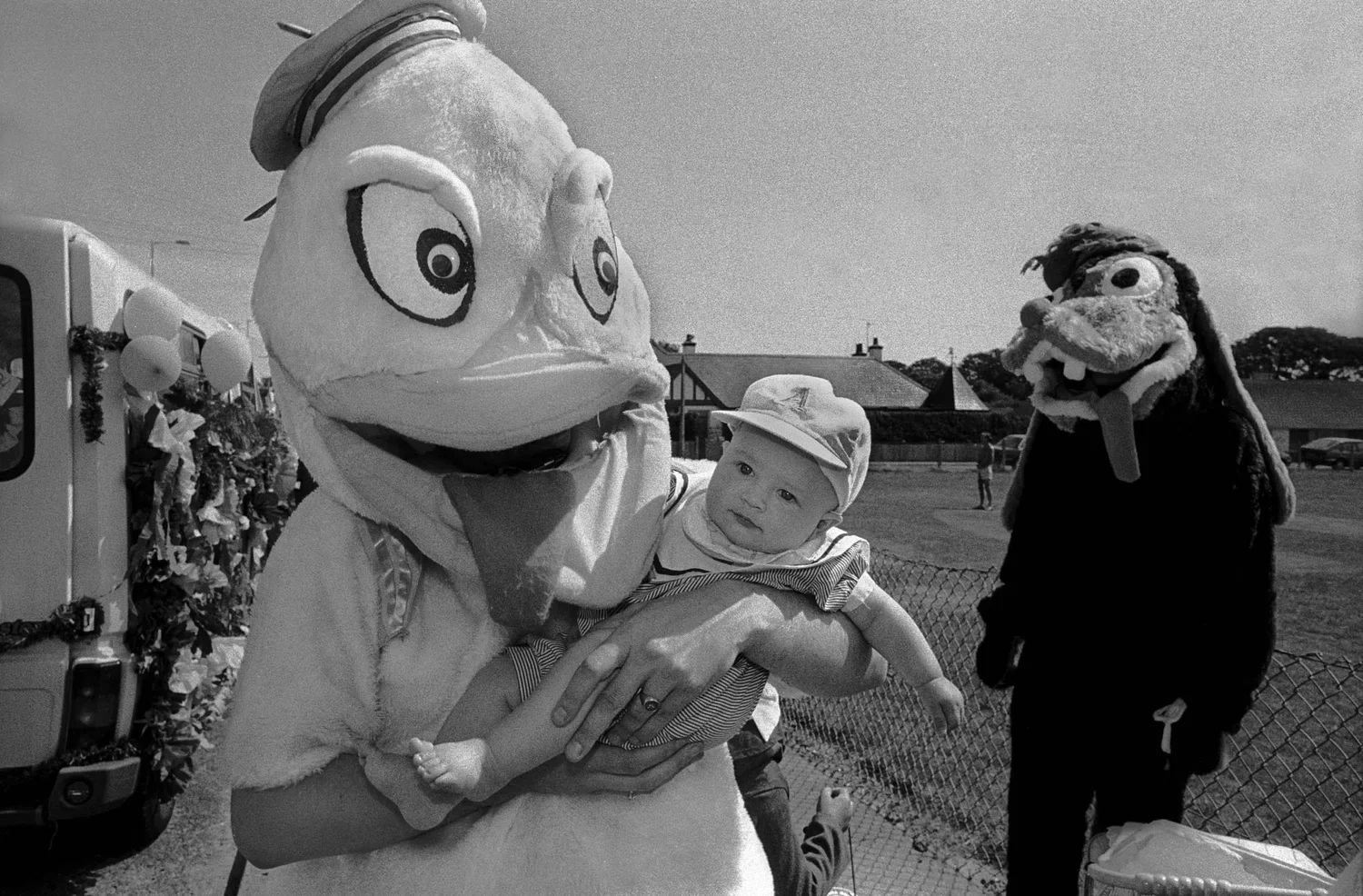 A child in a hat being held by a person in a mascot costume with a bird face at an outdoor event, with another mascot in a lion costume nearby.