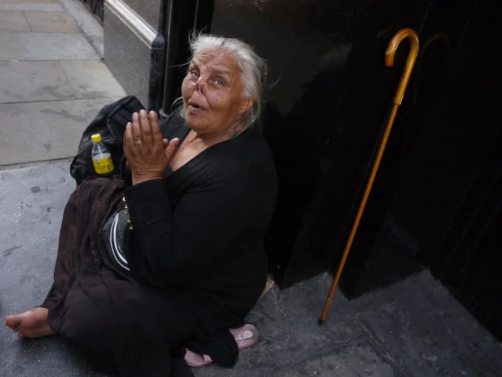 An elderly woman sitting on the sidewalk with hands clasped together, looking up with a distressed expression, next to a black wall and a wooden cane leaning against the wall.