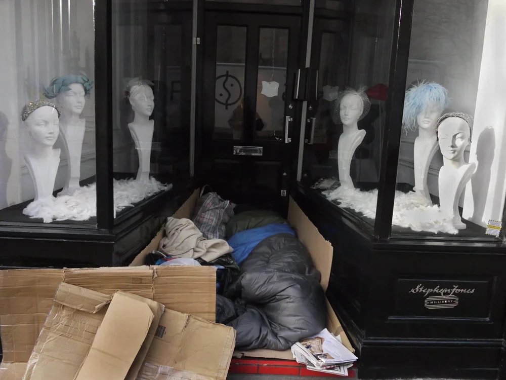 Disorganized area with mannequins displaying wigs behind glass in a storefront, cardboard boxes, a sleeping person on the floor, and scattered papers and clothes.