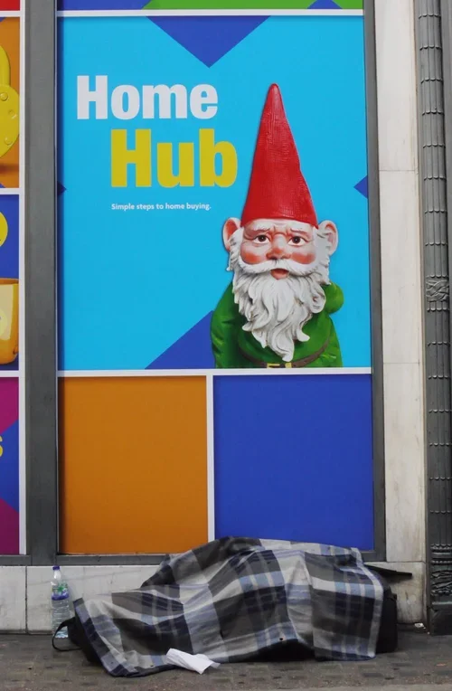 A colorful advertisement for Home Hub featuring a gnome with a red hat, white beard, and green outfit, alongside a person covered with a plaid blanket on the sidewalk.