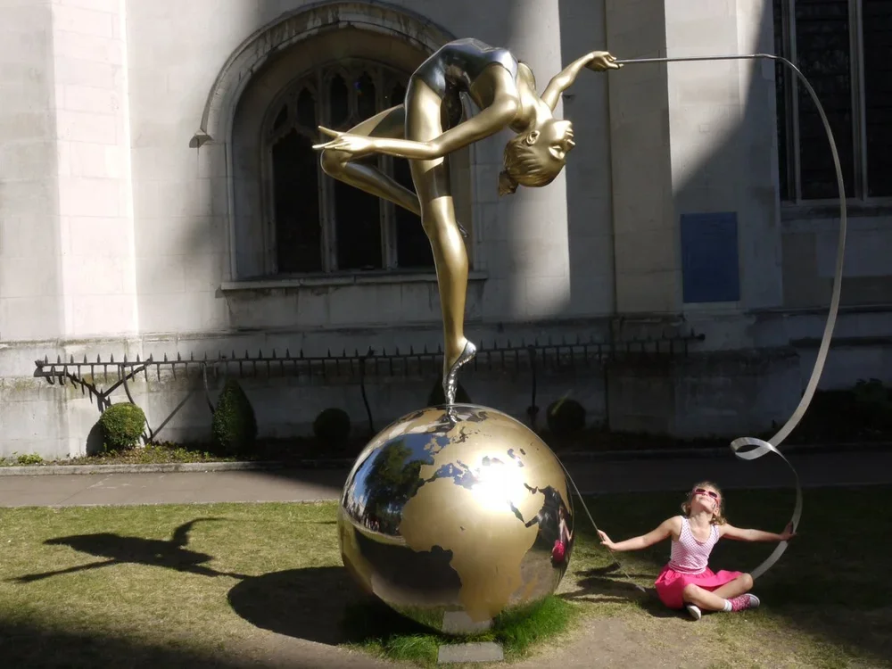 A girl wearing glasses and a pink skirt sitting on grass next to a large golden globe sculpture. The sculpture features a ballerina in a dance pose balancing on one leg on top of the globe, with her other leg extended behind her and holding a pole th