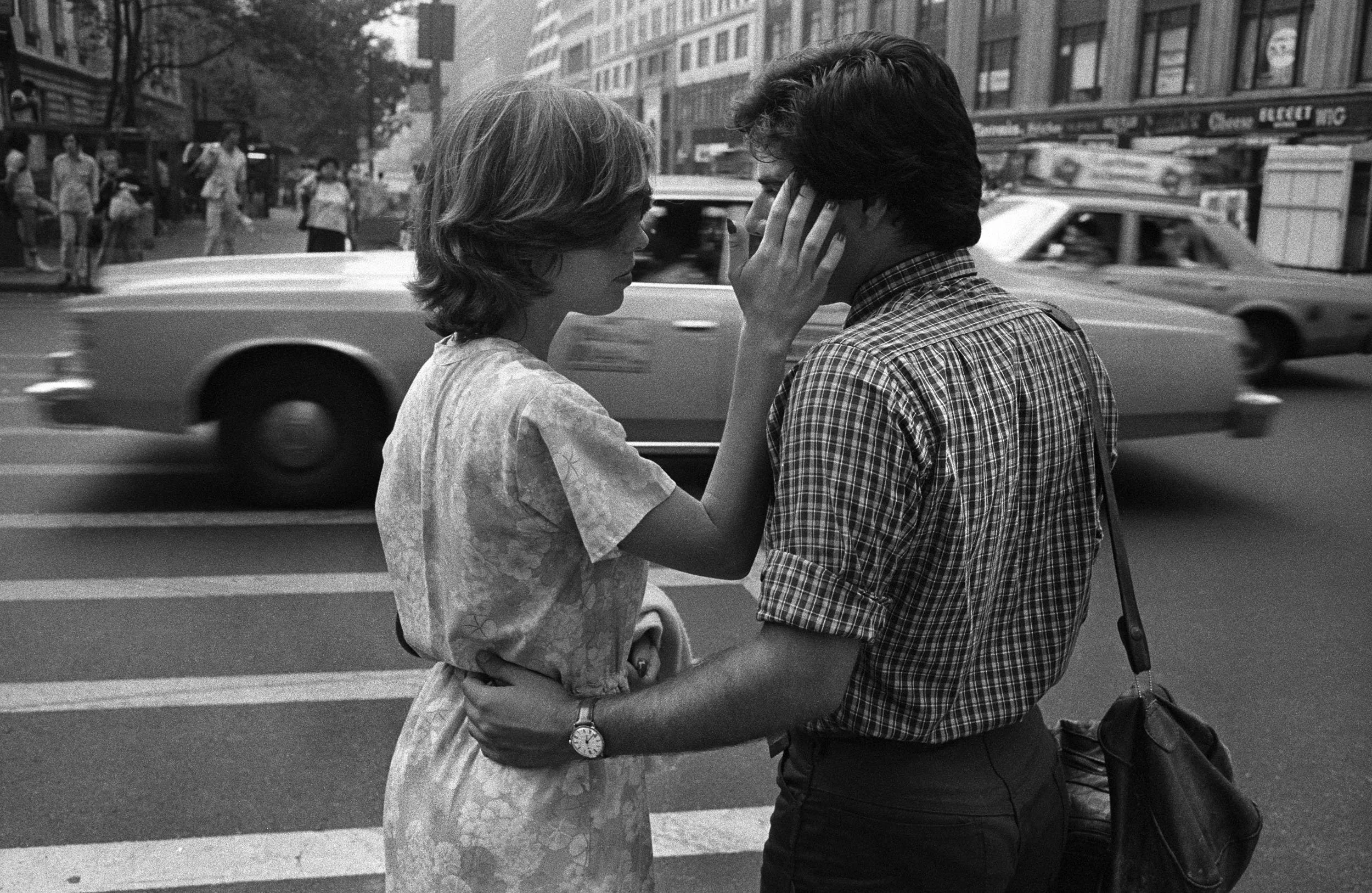 Two people, a woman and a man, are standing close to each other on a city street, touching foreheads with their eyes closed, conveying intimacy. The woman has short hair and is wearing a dress, while the man has dark hair and is wearing a checkered s