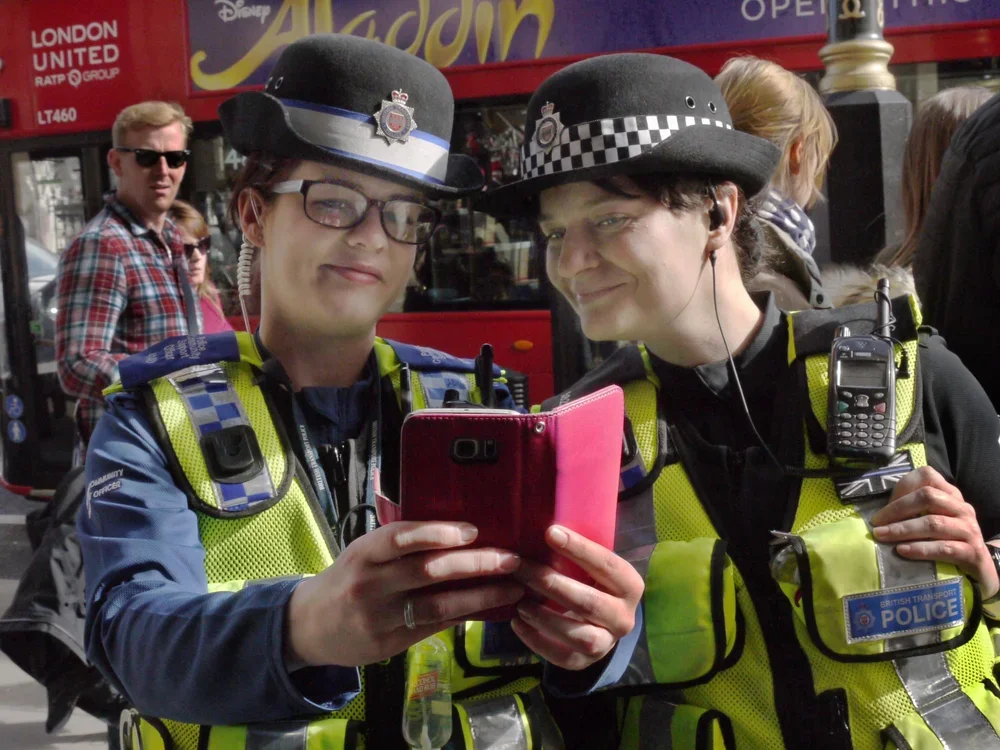 Two police officers, one male and one female, in uniform and high-visibility vests, are standing on a busy street, looking at a smartphone together. The female officer is holding the phone, and both are smiling. The background shows pedestrians and a