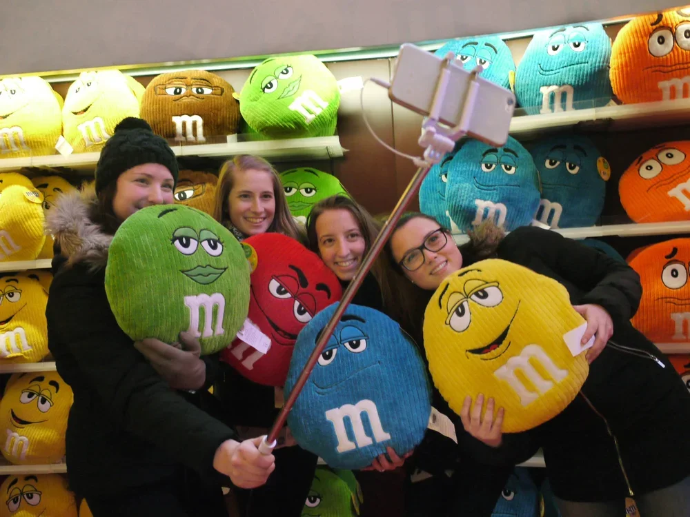 Group of four young women shopping for M&M's plush pillows in a store, holding and posing with colorful M&M's character pillows in yellow, green, red, and blue, with shelves of similar pillows in the background.