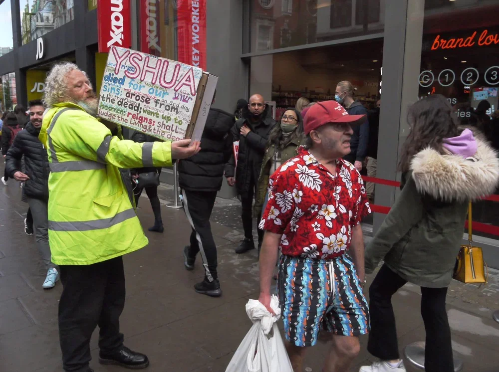 A man in a yellow safety vest holding a sign with colorful writing, standing on a busy city sidewalk. Several pedestrians are around, some wearing masks, with a storefront in the background.