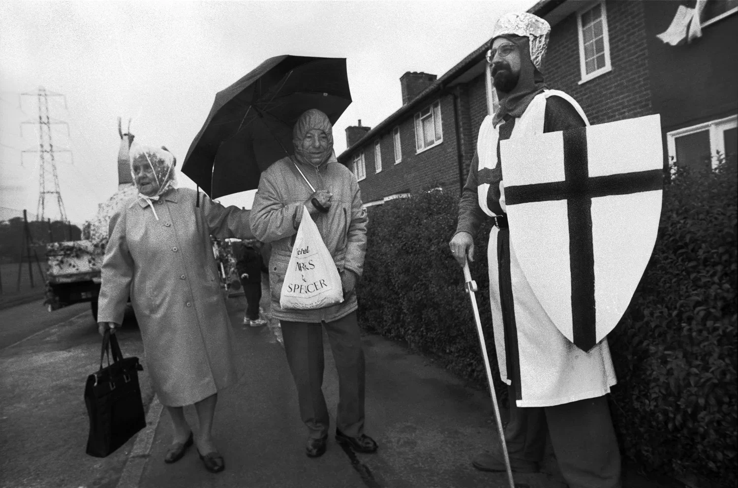 Two women and a man are walking on a sidewalk, with the man dressed as a medieval knight holding a shield. One woman holds an umbrella and wears a raincoat, while the other woman walks beside her carrying a bag. The man is wearing a costume with a wh