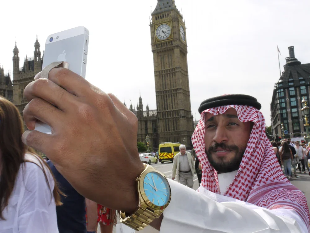 Man wearing a traditional Middle Eastern keffiyeh and a gold watch, taking a selfie with the Big Ben clock tower in London in the background, in a busy urban area.