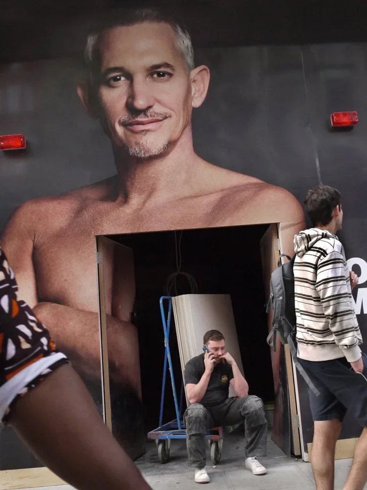 A large poster of a shirtless man with gray hair and a goatee on a dark wall. Three people are in front of the poster: one sitting on a small cart talking on a cellphone, and two walking by, one with a backpack and a striped hoodie.