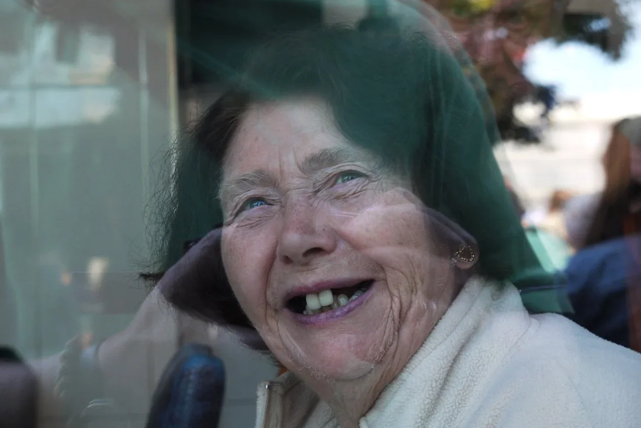 An elderly woman with gray hair, missing teeth, and a big smile, looking through a window.