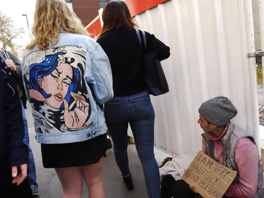 A woman holding a sign that says "I am very hungry. Good bless you," sitting on the sidewalk beside a woman in a black jacket and jeans. Two other women are standing nearby, one wearing a denim jacket with pop art design on the back. They appear to b