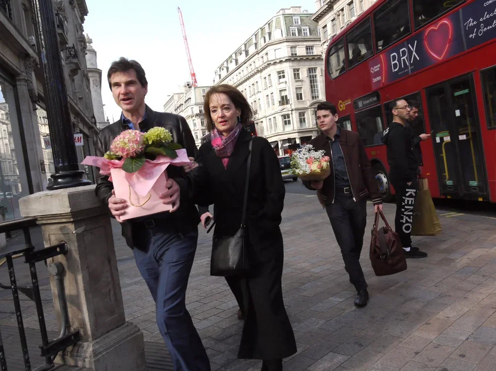 People walking on a city sidewalk next to a red double-decker bus, with buildings in the background. Two people in the foreground have flowers, and others are carrying bags.