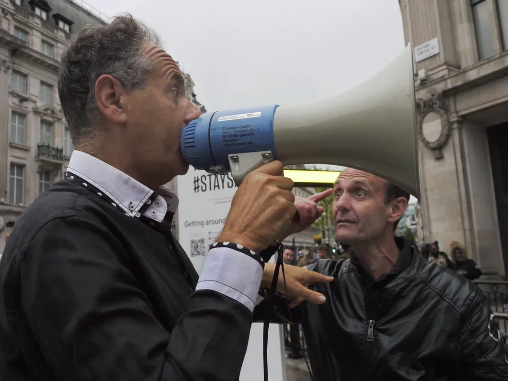 Two men are engaged in a confrontation on a city street; one man is shouting into a megaphone directed at the other man, who is pointing at him with an intense expression.
