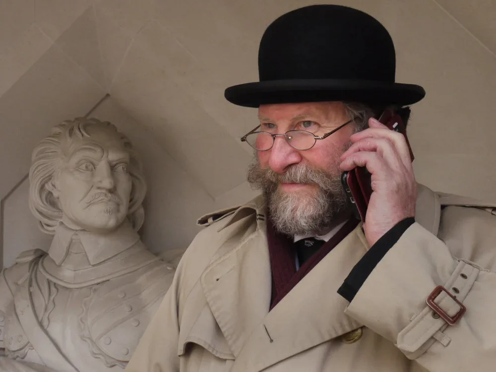 A man with a beard and glasses wearing a trench coat and bowler hat talking on a phone, standing next to a bust of a historical figure.