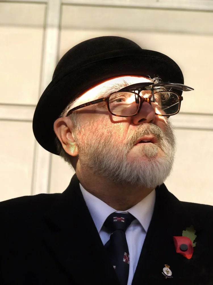 An older man with a beard and glasses, wearing a black bowler hat, a dark suit, white shirt, and tie, stands outside. The sunlight reflects on his glasses and hat.