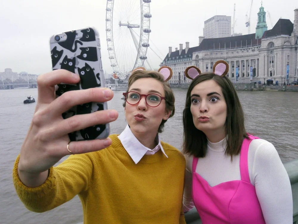 Two young women taking a selfie near the river, both wearing animal ear headbands, with the London Eye and historic buildings in the background.