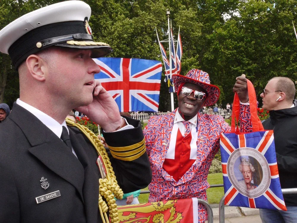 A man in a Royal Navy uniform with a name tag that reads 'De Savoye' talks on a cellphone, standing next to a man dressed in a colorful Union Jack patterned suit, red shirt, and matching hat, holding a tote with a photograph of Queen Elizabeth II, du