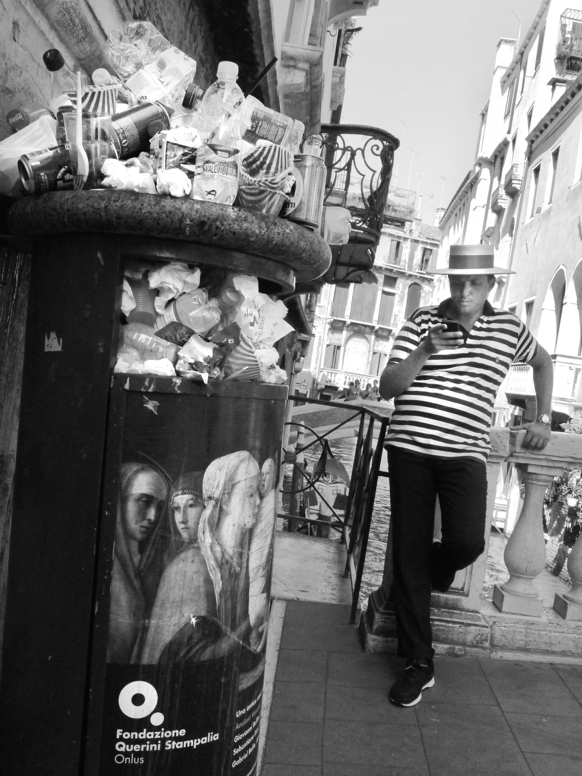 A young man in a striped shirt and wide-brimmed hat looks at his phone on a balcony in Venice, Italy. In the foreground, a trash or recycling bin features a poster with images of classical sculptures.