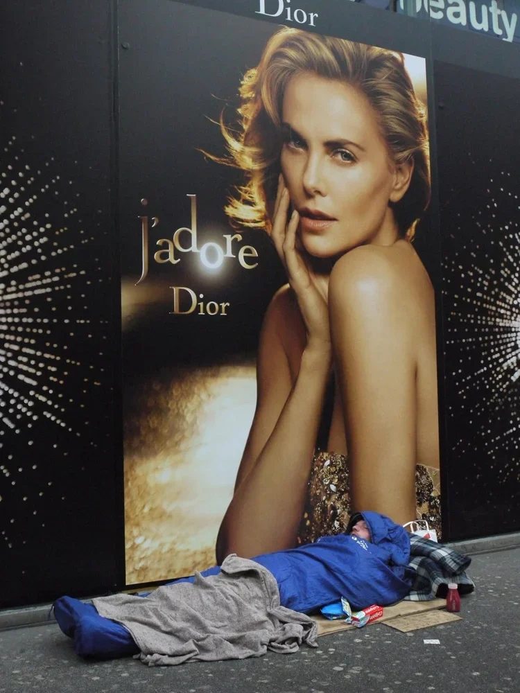 A homeless person sleeping on the ground in front of a large Dior advertisement featuring a woman with blonde hair and a glamorous pose.