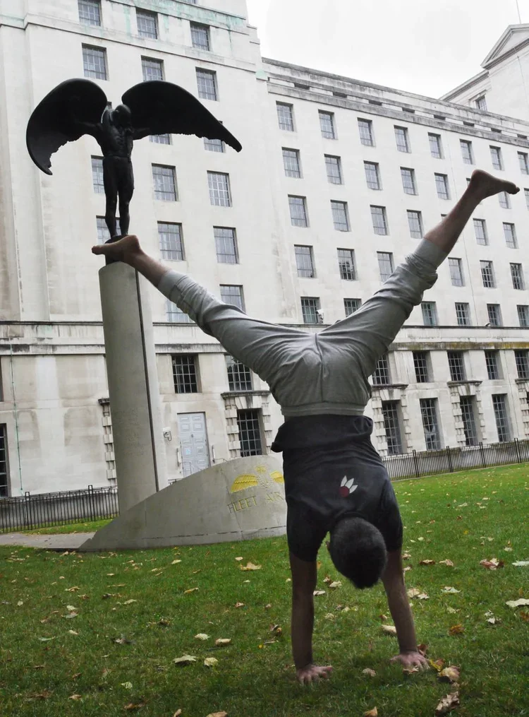 A person performing a handstand on the grass in front of a statue of an angel with wings on a tall pedestal, in an urban park area.