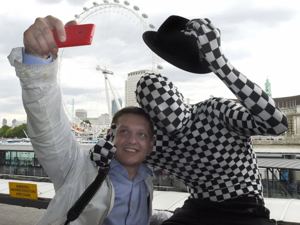 Two people taking a selfie in front of a London Eye ferris wheel. One person is wearing a white jacket and holding a red phone, while the other is dressed in a checkered outfit and black hat, adjusting their hat.