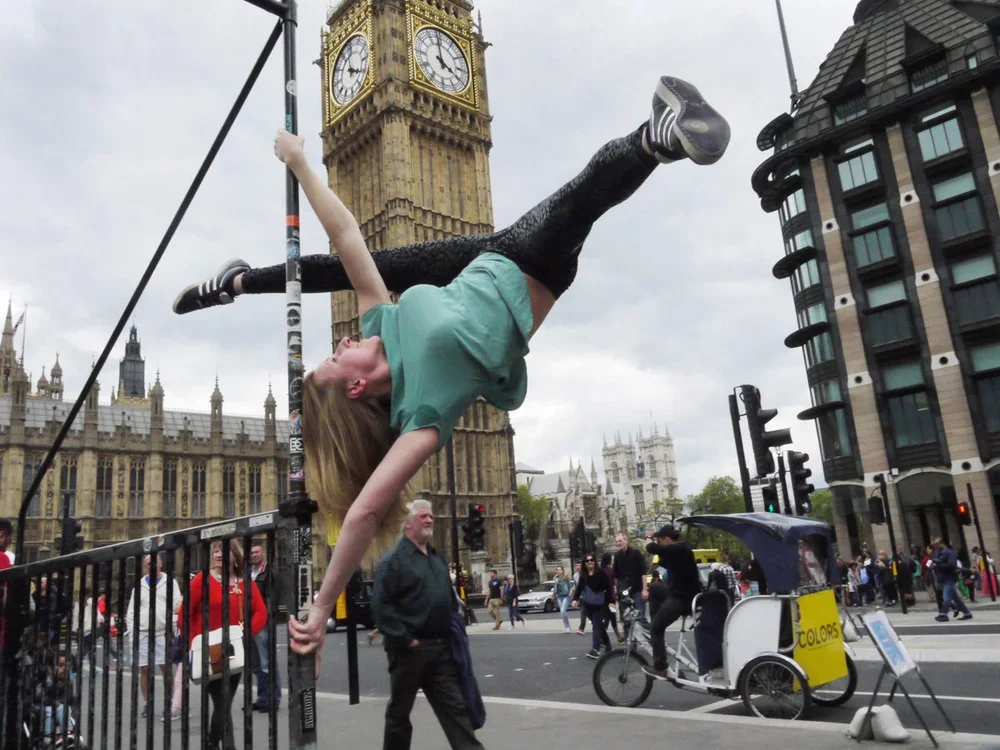 A woman performs on a circus unicycle in front of Big Ben in London, England, with her body upside down, hanging off the unicycle pole.