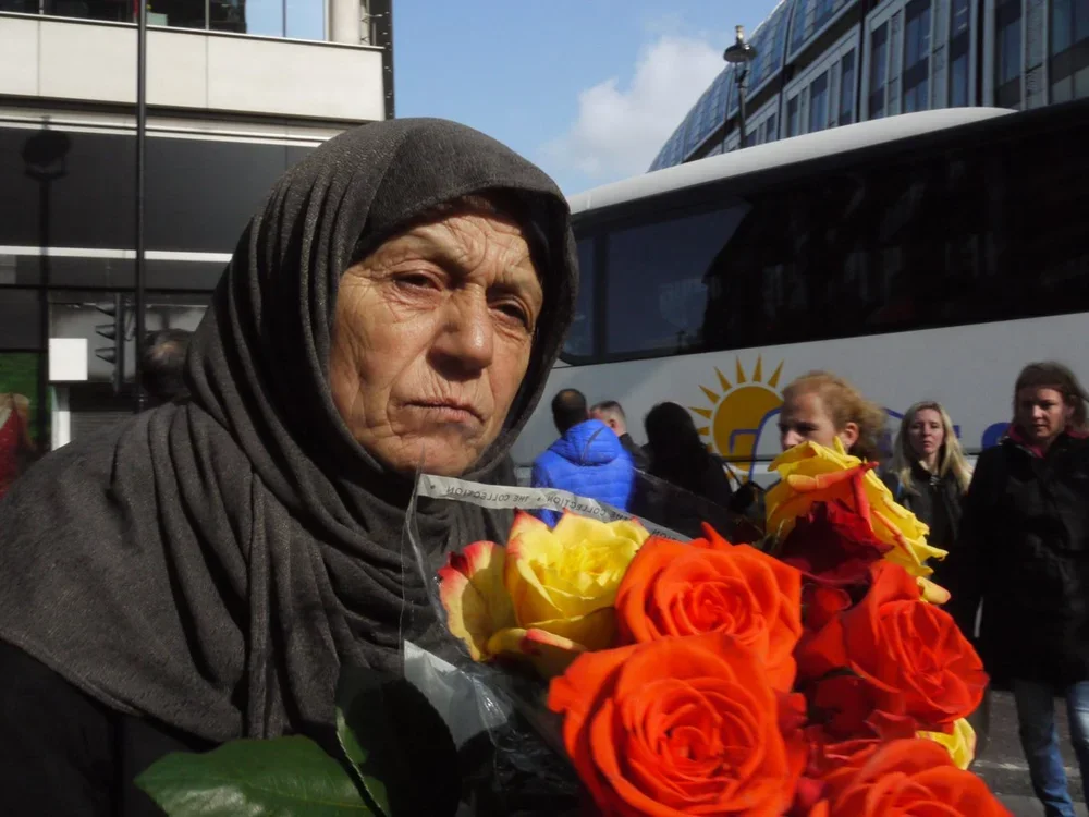 An older woman wearing a grey headscarf holding a bouquet of orange and yellow roses outdoors with a bus and several people in the background.