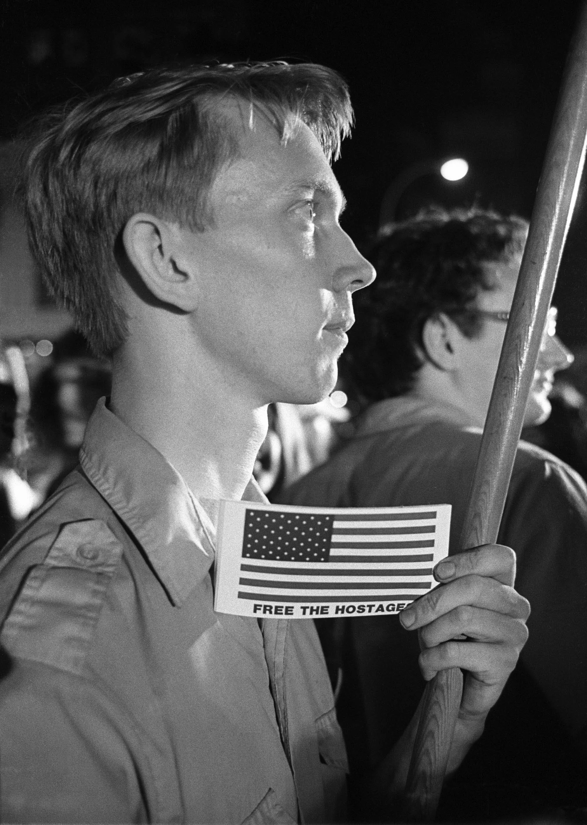 A young person with short hair holding a United States flag-shaped sign that reads 'Free the Hostages'. They are in a crowd with a dark background, and another person with sunglasses is partially visible behind them.