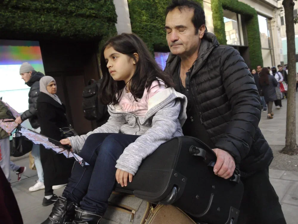 A man pushing a girl in a wheelchair on a city sidewalk, surrounded by pedestrians and trees.