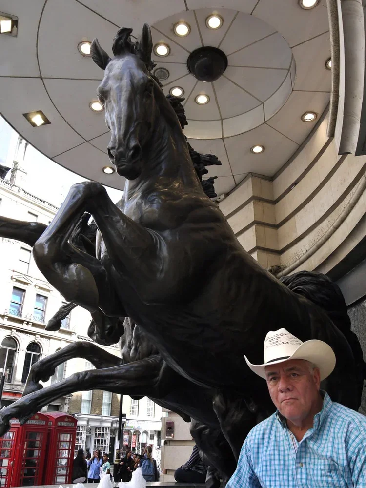 A man in a cowboy hat standing in front of a large black bronze statue of a horse rearing up on its hind legs, located outside a building with a curved ceiling and circular lighting fixtures.