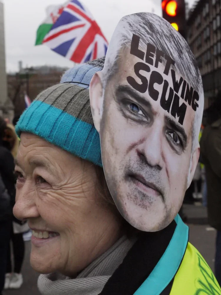 A person wearing a blue and gray striped beanie smiling at a protest, holding a mask of a man's face with the words "Left Winging Scum" written across the forehead.