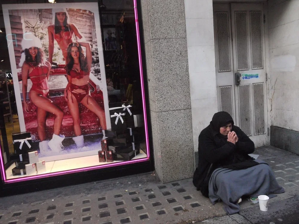 A woman dressed in a black hoodie and a grey skirt sits on the sidewalk with her hands clasped together in prayer or pleading. She is next to a cardboard cup and a small item on the ground. To her left, there is a store window displaying a large post