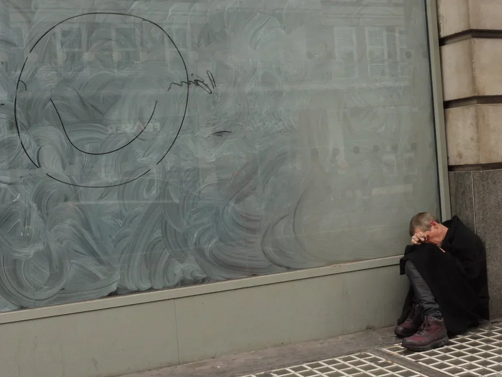 A man sitting on the sidewalk, leaning against a wall, with his head down and hand covering his face, next to a large foggy glass panel with a faint outline of a smiley face drawn on it.