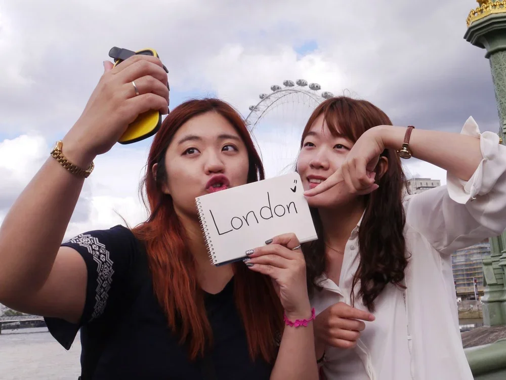 Two young women taking a selfie outdoors with a London Ferris wheel in the background, holding a sign that says 'London'.