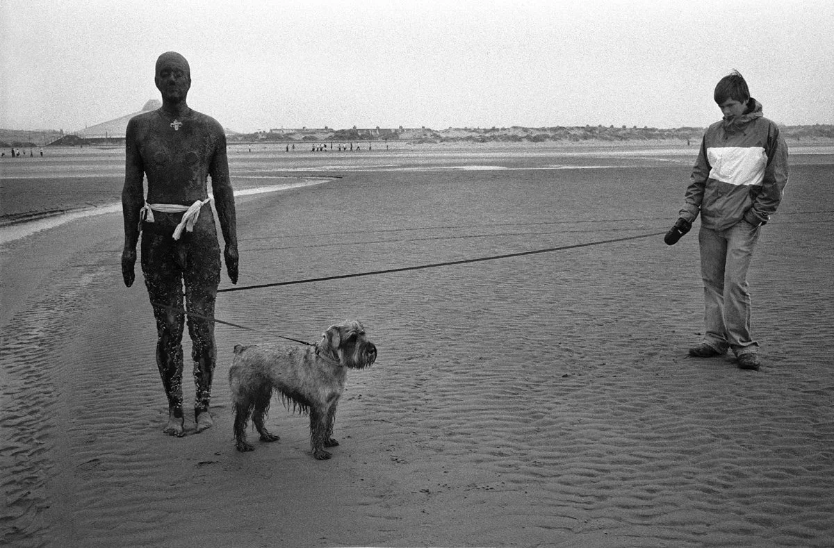 A black and white photo of a man with dark skin standing on a beach, holding a dog on a leash. The man has a towel around his waist and no shirt. A young boy dressed in a jacket and jeans stands nearby, holding the other end of the leash. The beach i