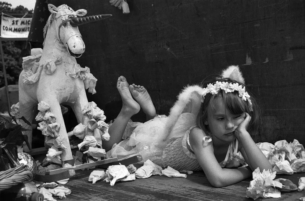 A young girl lying on the floor with a serious or bored expression, wearing a dress, floral headband, and cat ears, surrounded by torn paper. There is a unicorn stuffed animal nearby, and a dark wall in the background.