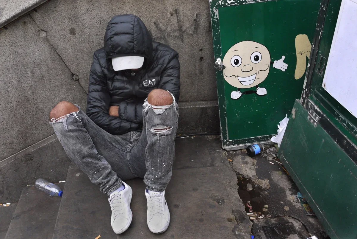 A person wearing a black hooded jacket and ripped jeans is sitting on the sidewalk with their head down and arms crossed, next to a decorated green box with a cartoon character face.