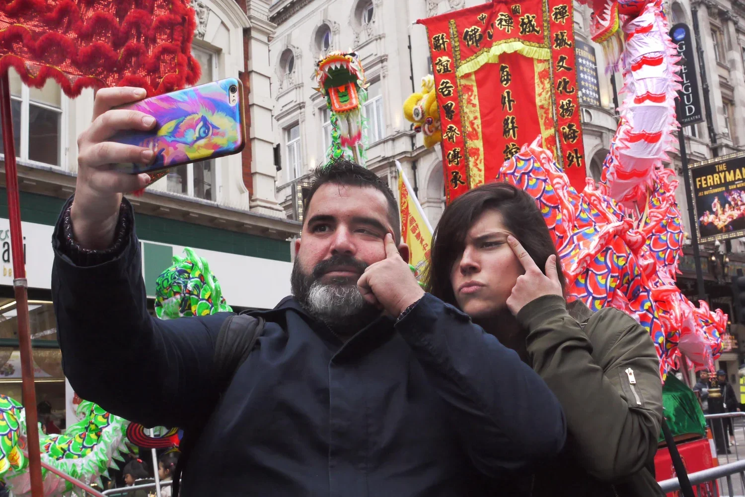 Two people taking a selfie at a Chinese New Year celebration in front of a colorful dragon parade float with red and yellow banners and decorations.