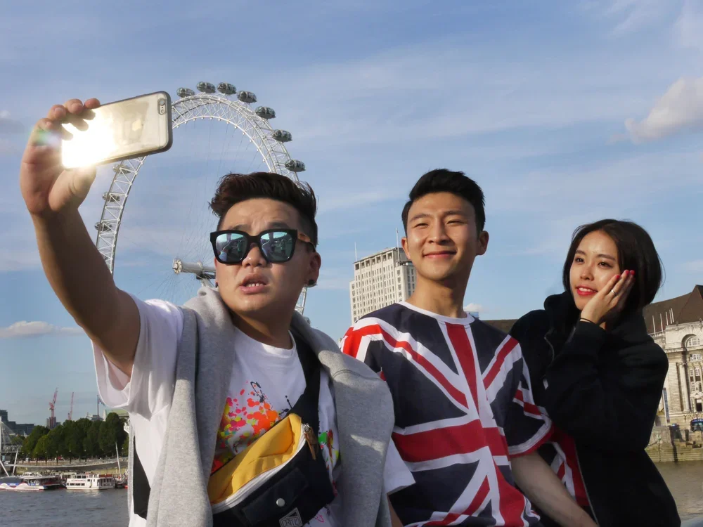 Three friends taking a selfie in front of a London Eye Ferris wheel, with a river and buildings in the background on a sunny day.