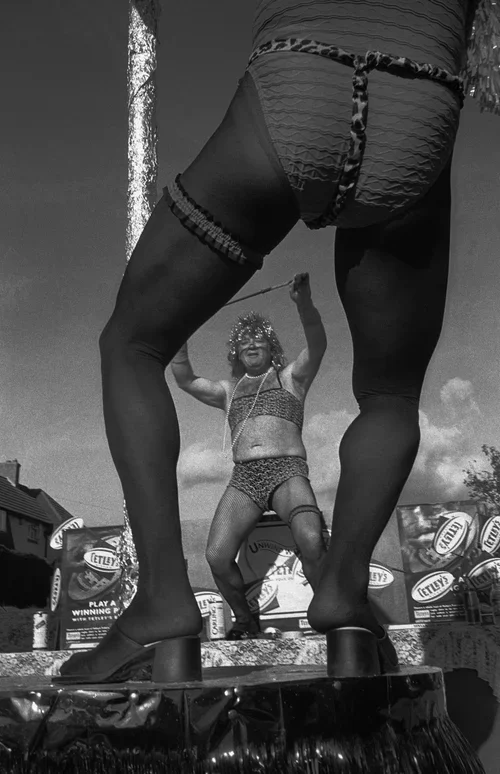 Woman dressed in lingerie holding a whip, seen from a low angle. A clown in a miniature costume and wig is in the background.