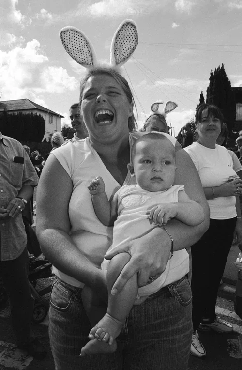A woman wearing bunny ears smiling and holding a baby, surrounded by people outdoors.