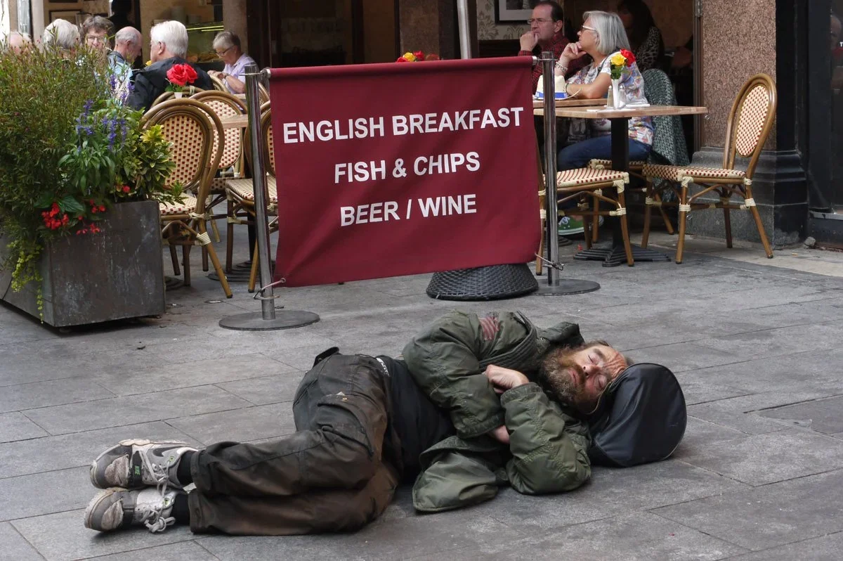 A man lying on the sidewalk next to a black backpack, sleeping or resting with a dark cap covering part of his face, outside a restaurant with outdoor seating. A large red sign in front of the restaurant advertises English breakfast, fish and chips, 