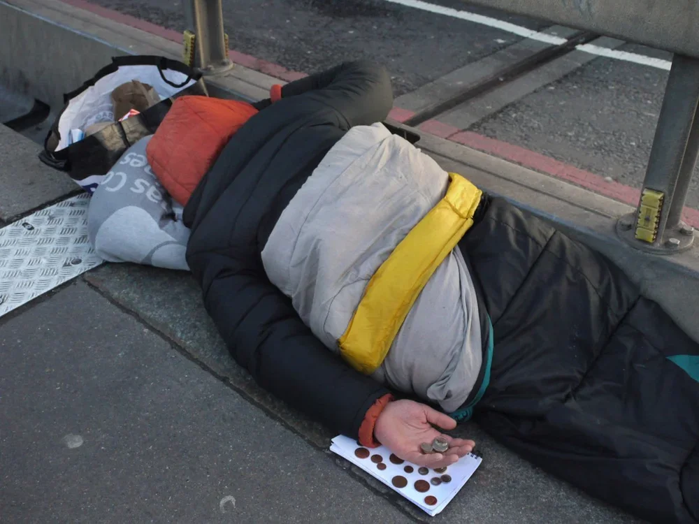 A person sleeping on the sidewalk using a makeshift mat, holding coins in one hand, beside some bags and belongings near a bus stop.