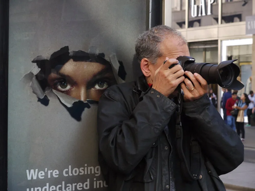 A man taking a photo with a Nikon camera next to a poster showing a woman's eyes and part of her face
